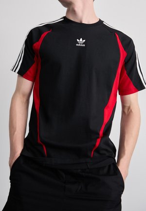 Mężczyzna w czarnej koszulce Adidas z czerwonymi bocznymi panelami i białymi paskami na rękawach na ramionach, ręce w kieszeniach, neutralne tło.