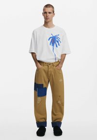 Vit oversized t-shirt med blå palmträdsgrafik, ihopparat med khakibyxor med olika lappar och slitna detaljer. Svart fotriktning.