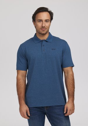 Mężczyzna w niebieskiej koszulce polo z krótkim rękawem i subtelnymi poziomymi paskami, stojący na jednolitym jasnym tle.