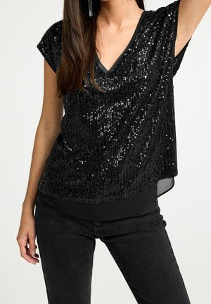 Woman wearing a black sequined short-sleeve top with V-neck and black pants, hand relaxed by side, hair down, wearing long earrings.