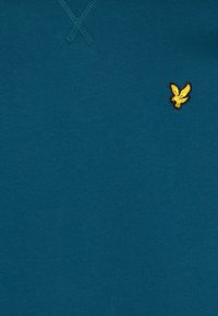 Tissu turquoise avec un petit logo de ronde jaune brodé sur le côté droit près du haut, et des détails de couture visibles au niveau de l'encolure.