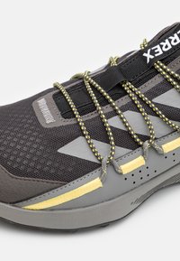 Scarpa sportiva grigia con materiali in mesh e sintetici, dotata di lacci con accenti gialli, superficie testurizzata e suola in gomma robusta con fori di drenaggio.