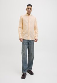 Lichtoranje button-up overhemd met een bandkraag, gecombineerd met recht toegenomen blauwe jeans en bruine schoenen, tegen een effen achtergrond.