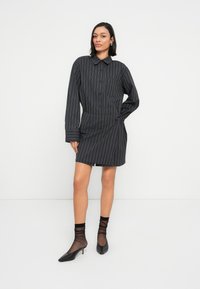 Μαύρο φόρεμα πουκάμισο με ρίγες, μακριά μανίκια, μπροστινά κουμπιά, λεπτομέρεια με τσέπη και πιέτες στο πλάι. Φορεμένο με διάφανα μαύρα καλτσάκια και μυτερές γόβες.