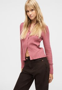 Femme blonde portant un cardigan rose tricoté boutonné et un pantalon marron foncé, se tenant légèrement appuyée contre un fond blanc uni.
