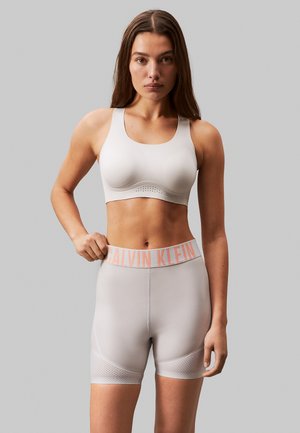 Femme en brassière de sport gris clair et short cycliste taille haute assorti avec une ceinture rose au logo Calvin Klein, debout devant un fond gris uni.