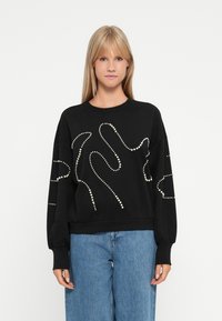 Sweat-shirt noir en tissu texturé, avec des accents blancs en forme de perles dans des motifs abstraits. Coupe oversize et manches longues.