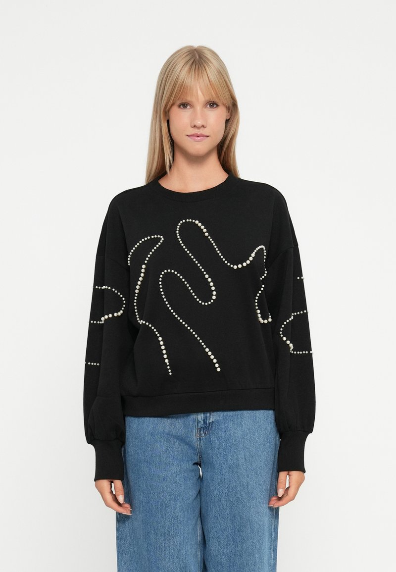 Sweat-shirt noir en tissu texturé, avec des accents blancs en forme de perles dans des motifs abstraits. Coupe oversize et manches longues.