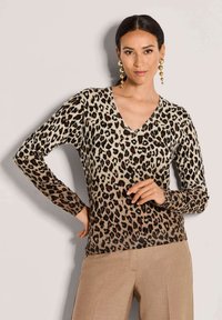 Frau mit dunklem Haar, die goldene Anhänger-Ohrringe, einen V-Ausschnitt Pullover im Leopardenmuster und beige Hosen trägt, vor einem neutralen Hintergrund.