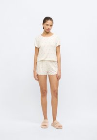 Ελαφρύ σετ πιτζάμας κρέμας με μικρά ροζ μοτίβα καρδιών. Μπλούζα με κοντά μανίκια και σορτς με κυματιστό τελείωμα. Συνδυασμένο με μπεζ χνουδωτές παντόφλες.