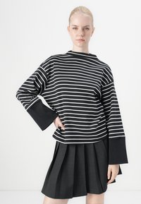 Blusa de mangas longas às riscas pretas e brancas com mangas largas, combinada com uma saia plissada preta, destacando a textura e cores contrastantes.