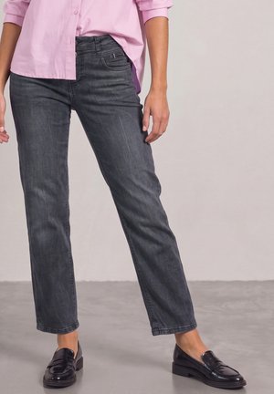 Femme portant un jean droit gris foncé, une chemise rose clair boutonnée et des mocassins noirs, debout sur un sol gris.
