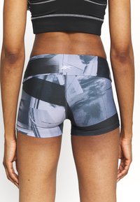Femme portant un short de sport à motif abstrait noir et gris et un brassard de sport noir, vue de dos sur un fond blanc.