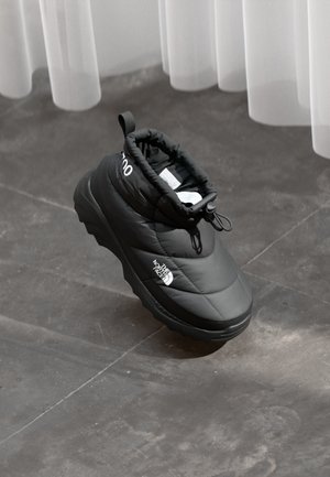 Czarny ocieplany but wsuwany North Face z grubą podeszwą, spoczywający na szarej kafelkowej podłodze pod przezroczystymi białymi zasłonami.