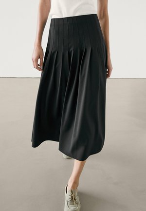 Femme marchant portant une jupe midi plissée noire, un haut blanc et des chaussures à lacets gris clair sur un sol uni.