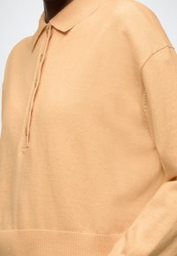 Persiku krāsas džemperis ar apkakli, trīs pogu rāvēju, ribotām malām un gludu faktūru.