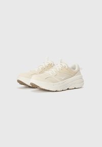 Beige sneakers with textured mesh upper, white accents, cushioned sole, and speckled detailing on the outsole. Laces secured with loops.
