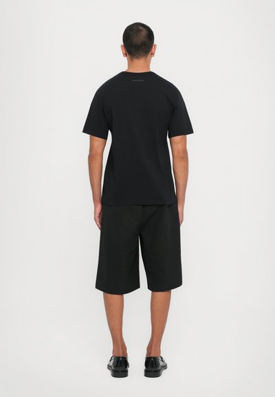 T-shirt en coton noir à manches courtes, associé à un short noir jusqu'aux genoux, porté avec des chaussures noires. Design minimaliste, sans motifs visibles.