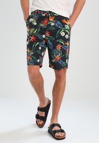 Uomo che indossa pantaloni corti neri a fiori e sandali neri con fibbia, con le mani nelle tasche, che cammina su un pavimento bianco.