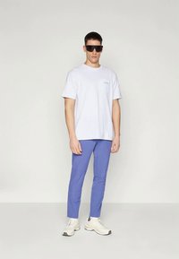 Balts īsām piedurknēm t-krekls ar mazu logotipu, apvienots ar vienkāršiem ziliem biksēm un baltām kedām; indivīds valkā lielas melnas saulesbrilles.