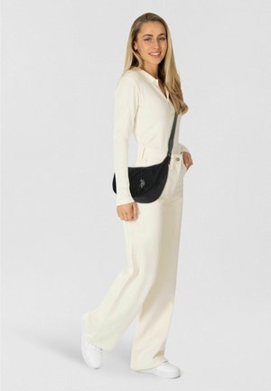 Zwarte schoudertas met een gebogen vorm, voorzien van een gestructureerd oppervlak en een designelement in het midden. Model draagt kleding in lichte kleuren.