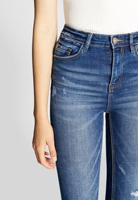 Kvinna i midjehöga blå skinny jeans med blekta och slitna detaljer, tillsammans med en vit ribbad ärmlös topp.
