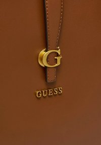 Borsa in pelle marrone con logo dorato "G" e testo "GUESS" in rilievo, caratterizzata da una texture liscia e un design semplice.