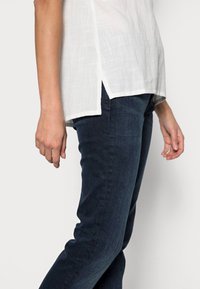 Biała lniana bluzka o teksturowanym wykończeniu i bocznych rozcięciach, zestawiona z ciemnoniebieskimi dżinsami w wąskim kroju. Skupienie na kontraście tkaniny i kolorów.