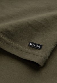 Olivenfarbener T-Shirt-Stoff mit glatter Textur. Verfügt über einen gefalteten Rand und ein schwarzes Etikett mit "ombre" in weißen Buchstaben.