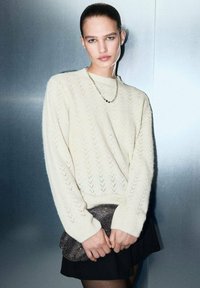 Pull crème texturé avec un motif en chevrons, associé à une jupe sombre et tenant une pochette métallique. L'arrière-plan est une surface lisse en argent.