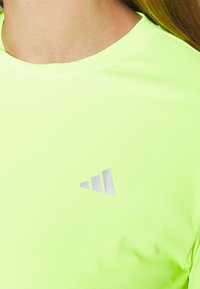 Fluorescerande gul idrottströja gjord av slätt, elastiskt material, med en reflexgrå logotyp på övre vänstra sidan.