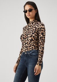 Mujer que lleva gafas de sol de estilo cat-eye negro, una blusa de manga larga con estampado de leopardo y jeans azul oscuro, de pie contra un fondo liso claro.