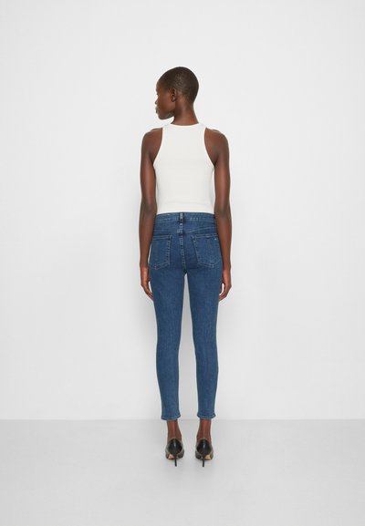 Calças de ganga escuras de cintura alta, com um design justo, dois bolsos traseiros e um ligeiro estiramento. Combinadas com uma blusa branca sem mangas e sapatos de salto pretos.