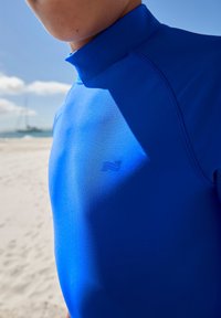 Modra potapljaška obleka z visokim ovratnikom, gladke teksture in subtilnim logotipom na prsih. Brezšivna zasnova z kontrastnimi šivi.