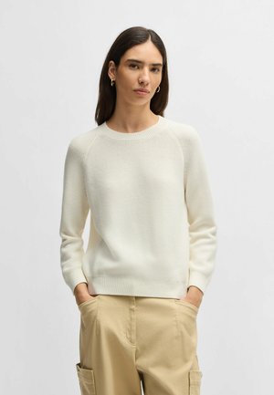 Light cream ribbed knit sweater with a crew neckline, raglan sleeves, and a fitted hem. Worn with beige cargo pants, featured pockets.