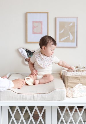 Baby i en blomstrete smekke som sitter på en stripete stellematte, rekker mot en beige blomstrete organizer, med to abstrakte innrammede kunstverk på veggen.