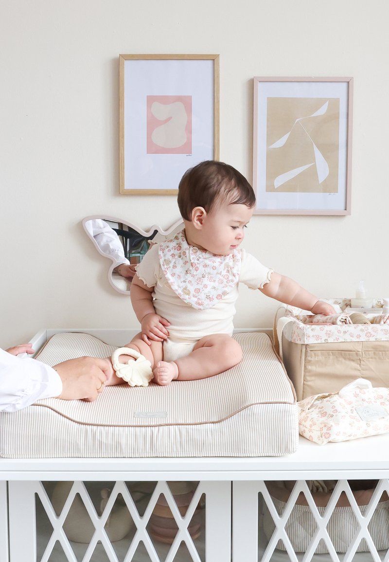 Bambino con bavaglino floreale seduto su un cuscino fasciatoio a righe, che afferra un organizzatore floreale beige, con due opere d'arte astratte incorniciate sulla parete.