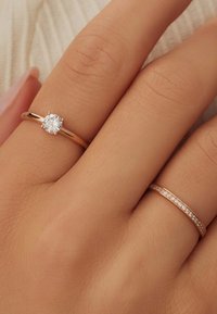 Goldene Verlobungsring mit einem runden, klaren Edelstein, begleitet von einem dünnen goldenen Band mit kleinen eingearbeiteten Steinen, auf einer Hand mit neutralem Hautton.