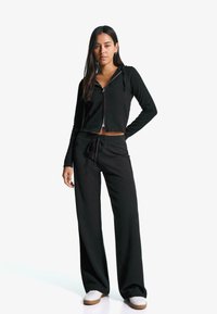 Čierna skrátená zipsová mikina s dlhými rukávmi, doplnená o čierne nohavice s vysokým pásom a sypeťom. Model má na sebe biele tenisky.