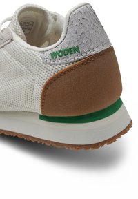 Baskets en maille blanche avec talon gris texturé, accents marron et vert, et col rembourré portant le mot "WOODEN" en broderie verte. Semelle extérieure en caoutchouc.