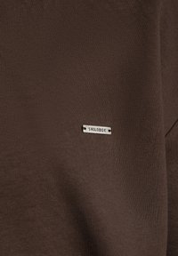 Tissu marron avec une petite étiquette métallique rectangulaire cousue, gravée du mot "SMILODOX" en lettres majuscules.