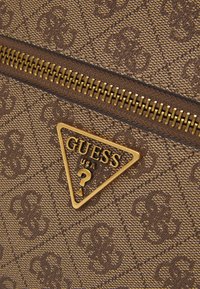 Borsa a tracolla in tessuto marrone con motivo, con una cerniera dorata e un'etichetta a forma di triangolo con la scritta 'GUESS USA'. Materiale testurizzato con disegni sottili.