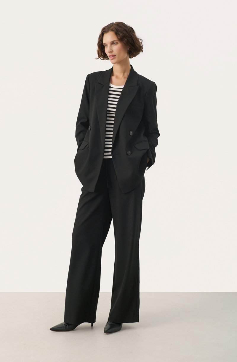 Schwarzer doppelt geknöpfter Blazer mit zwei vorderen Taschen, kombiniert mit schwarzen weit geschnittenen Hosen. Darunter trägt die Person ein schwarz-weiß gestreiftes Hemd.