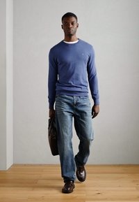 Blauer langärmeliger Pullover, hellblaue Jeans, braune Schuhe, hält eine braune Ledertasche, steht auf einem Holzfußboden vor einem neutralen Hintergrund.