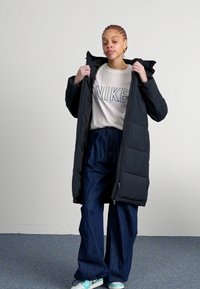 Zwarte pufferjas met een capuchon, met een gequilt ontwerp en verticale naden. Gekoopt met een lichtgrijze Nike-logo shirt en blauwe wijde jeans.