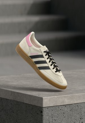 Άσπρο και ροζ αθλητικό παπούτσι Adidas με μαύρες ρίγες και καφέ σόλα, ισορροπώντας σε γκρι τσιμεντένια άκρη με θολά σκαλοπάτια στο φόντο.