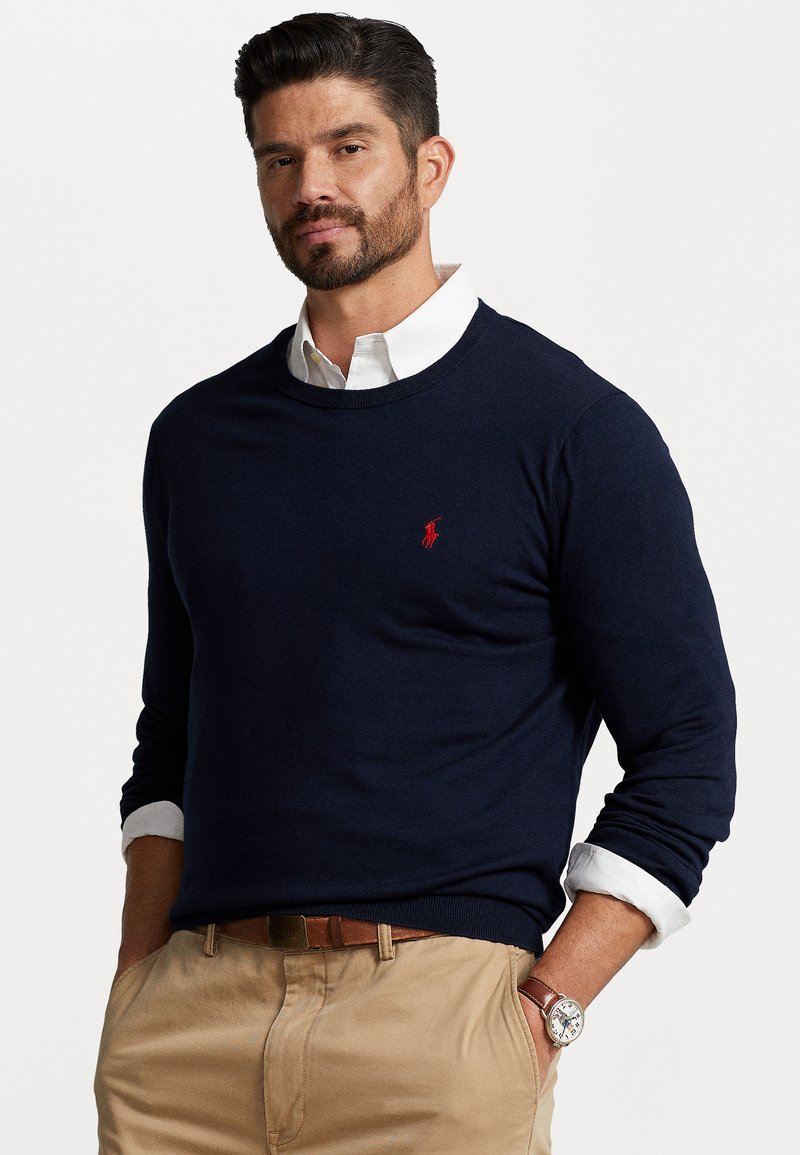 Suéter de cuello redondo azul marino hecho de un material de punto suave, con un pequeño logotipo rojo bordado en el pecho. Mangas enrolladas.