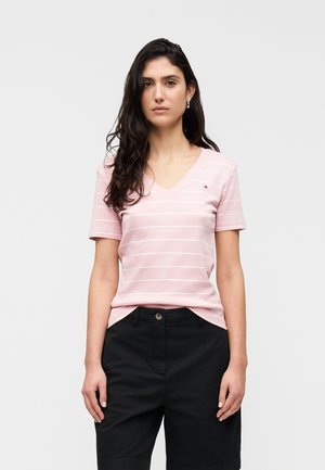 Frau steht, trägt ein hellrosa V-Ausschnitt kurzärmeliges gestreiftes Shirt und schwarze hochtaillierte Hose, schaut nach vorne mit entspannten Armen.