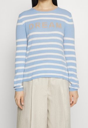 Personne portant un pull à rayures bleu clair et blanc avec l’inscription « DREAM » en beige, associé à un pantalon large beige clair.