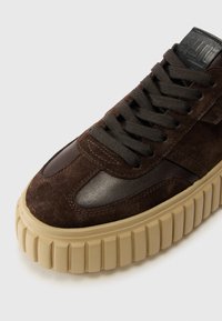 Sneakers en suede marron avec des accents en cuir, semelle en caoutchouc épaisse et lacets texturés. Présente un bout arrondi et une languette rembourrée pour le confort.